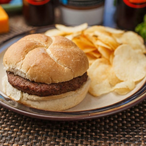A charbroiled steak burger on a bun with a side of potato chips on a plate.